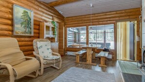 En stuga byggd av timmer, med träväggar, en matplats med bänkar, en öppen spis och ett stort fönster med utsikt över träden.