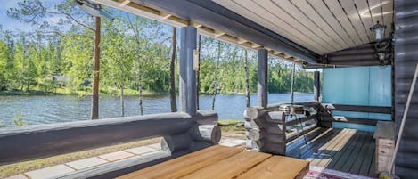 An outdoor wooden deck with benches and a grill, overlooking a lake with trees and a distant building.