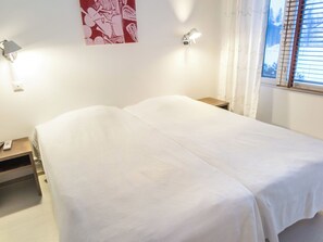 Cama com colcha branca em quarto com parede branca, mesa de cabeceira pequena e luminária fixada na parede.