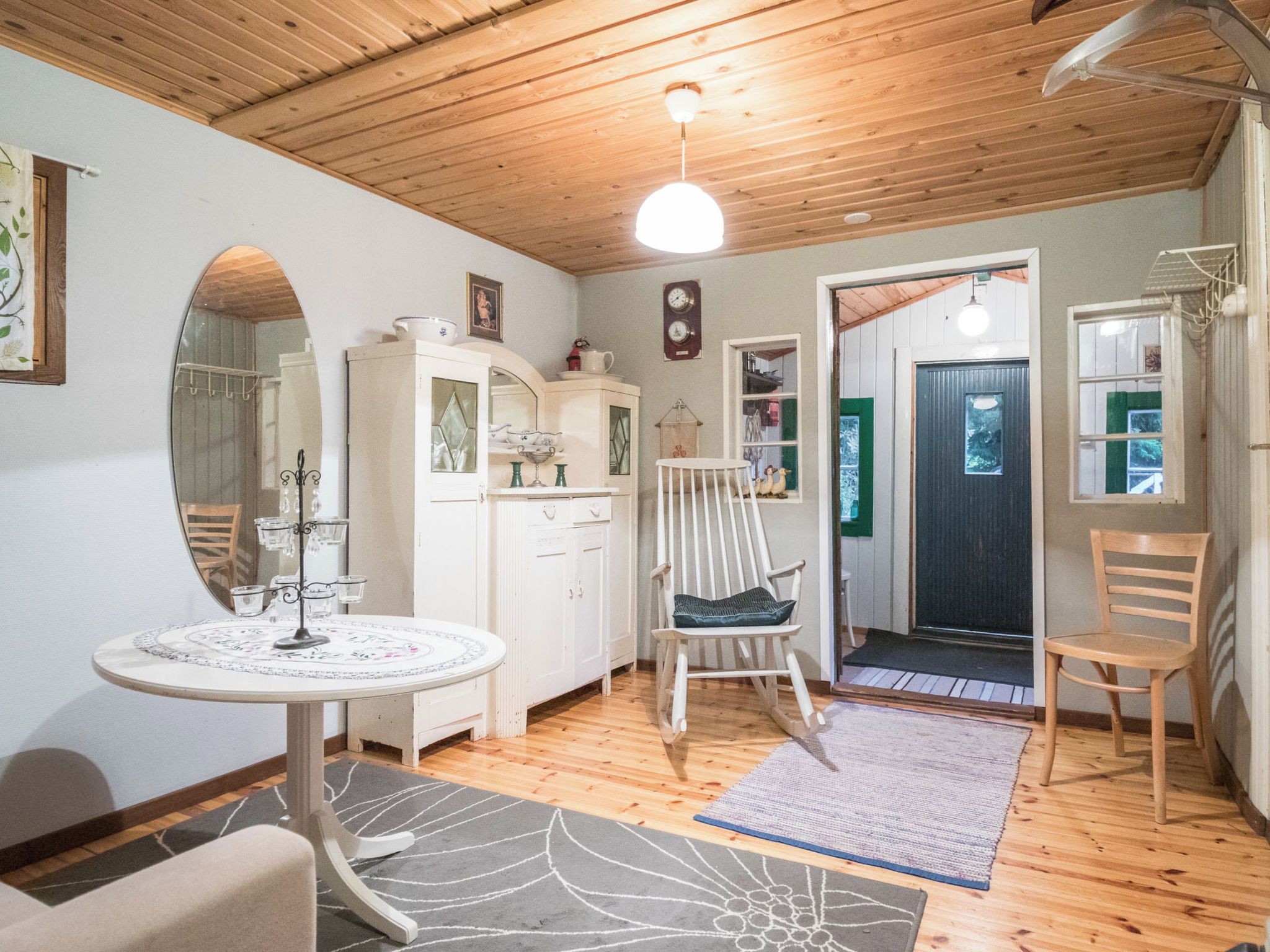 Una habitación acogedora con techo de madera, una mesa redonda con mantel floral, un aparador blanco con puertas de cristal, un sillón mecedora y una puerta que da al exterior.