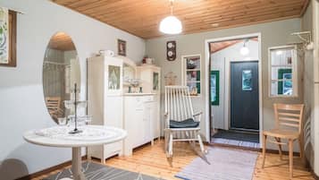 Una stanza accogliente con soffitto in legno, un tavolino rotondo con tovaglia floreale, un armadietto bianco con vetri, una poltrona a dondolo e una porta che dà sull'esterno.