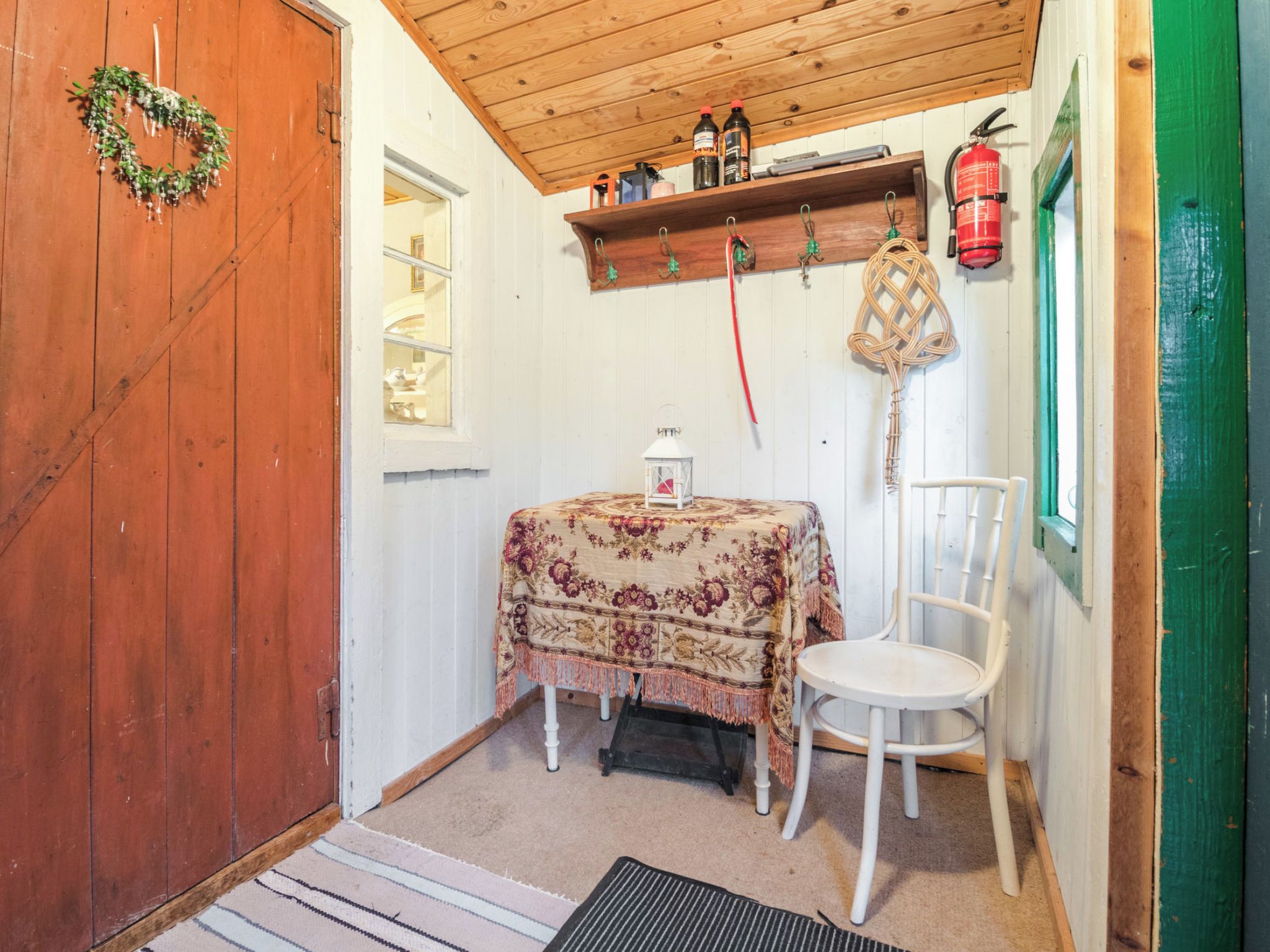 Habitació amb una porta de fusta, taula amb camavisor floral, cadira blanca, prestatge amb ampolles, fanal i extintor.