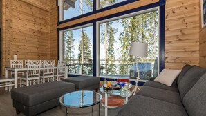 A cozy cabin interior with a large window, a sofa, a glass coffee table, and a dining area with chairs.