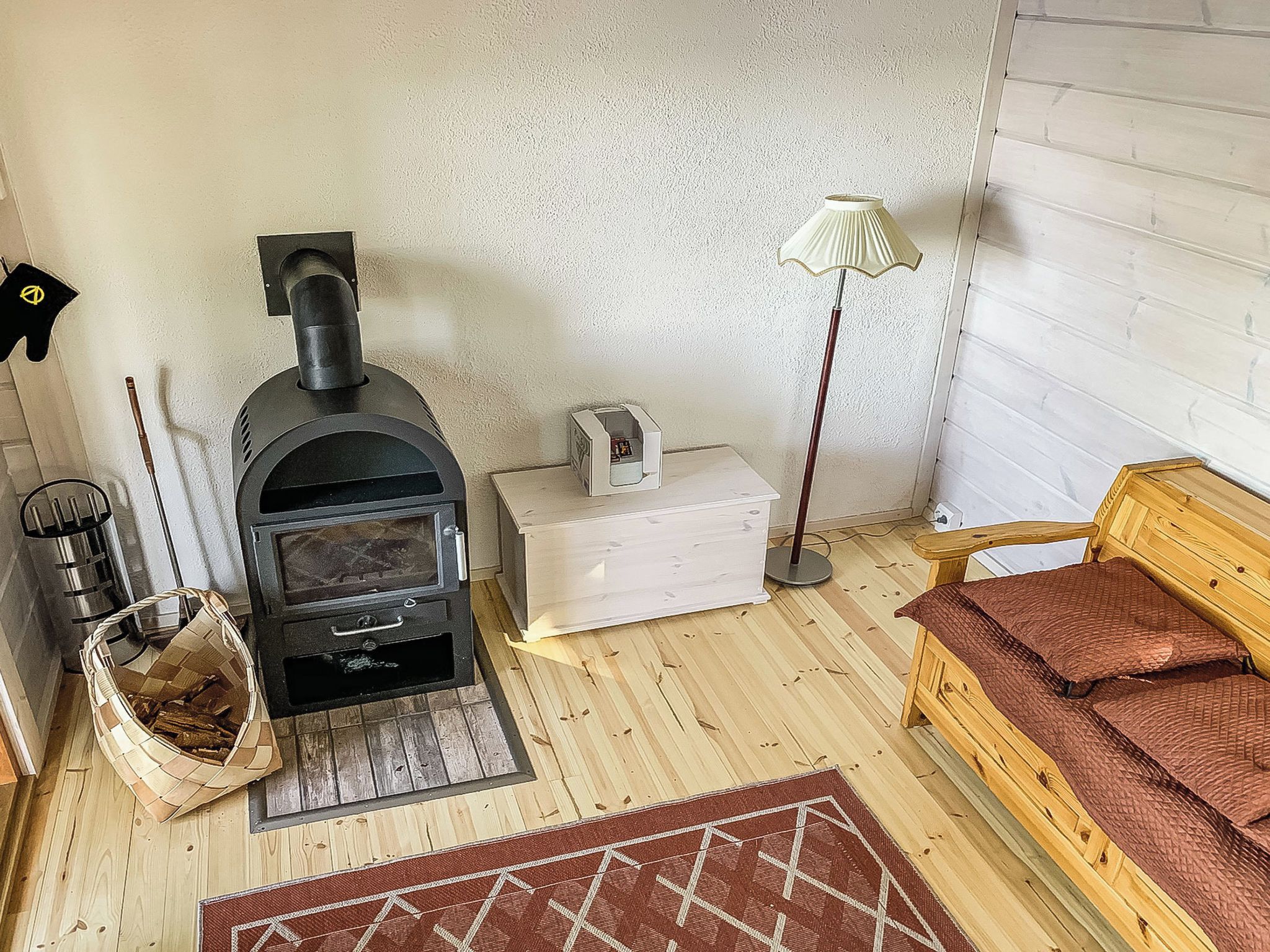 Una habitación acogedora con una estufa de leña, un caja de almacenamiento blanco, una lámpara, una cesta tejida y un banco de madera.
