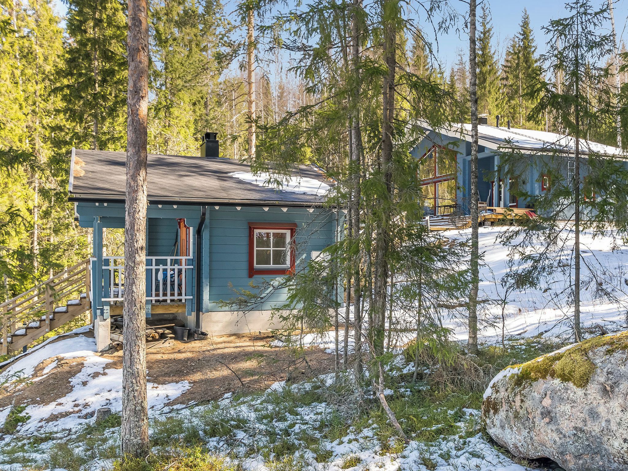 Mökki, jossa on kuisti, ja sen ympärillä on puita ja lunta.