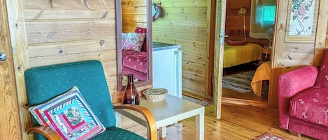 A cozy cabin interior with a wooden wall, a teal armchair with a patterned pillow, a small table, a red sofa, and a wooden floor.