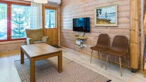 Ein gemütliches Blockhaus-Interieur mit Holzausstattung, einem an der Wand montierten Fernseher und einem großen Fenster mit Blick auf Bäume.