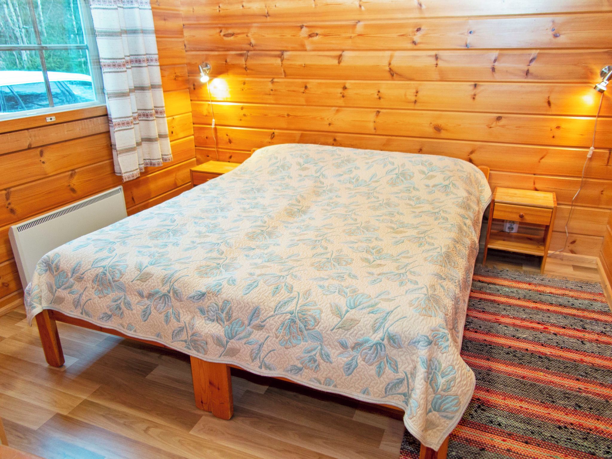 A wooden cabin room with a bed, two bedside tables, a heater, and a window with a view of trees.