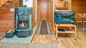 A cozy cabin interior with a stone fireplace, wooden walls, a wooden staircase, a dining area, and a television displaying a mountain landscape. - Rinnepelto by Interhome (Nilsiä)