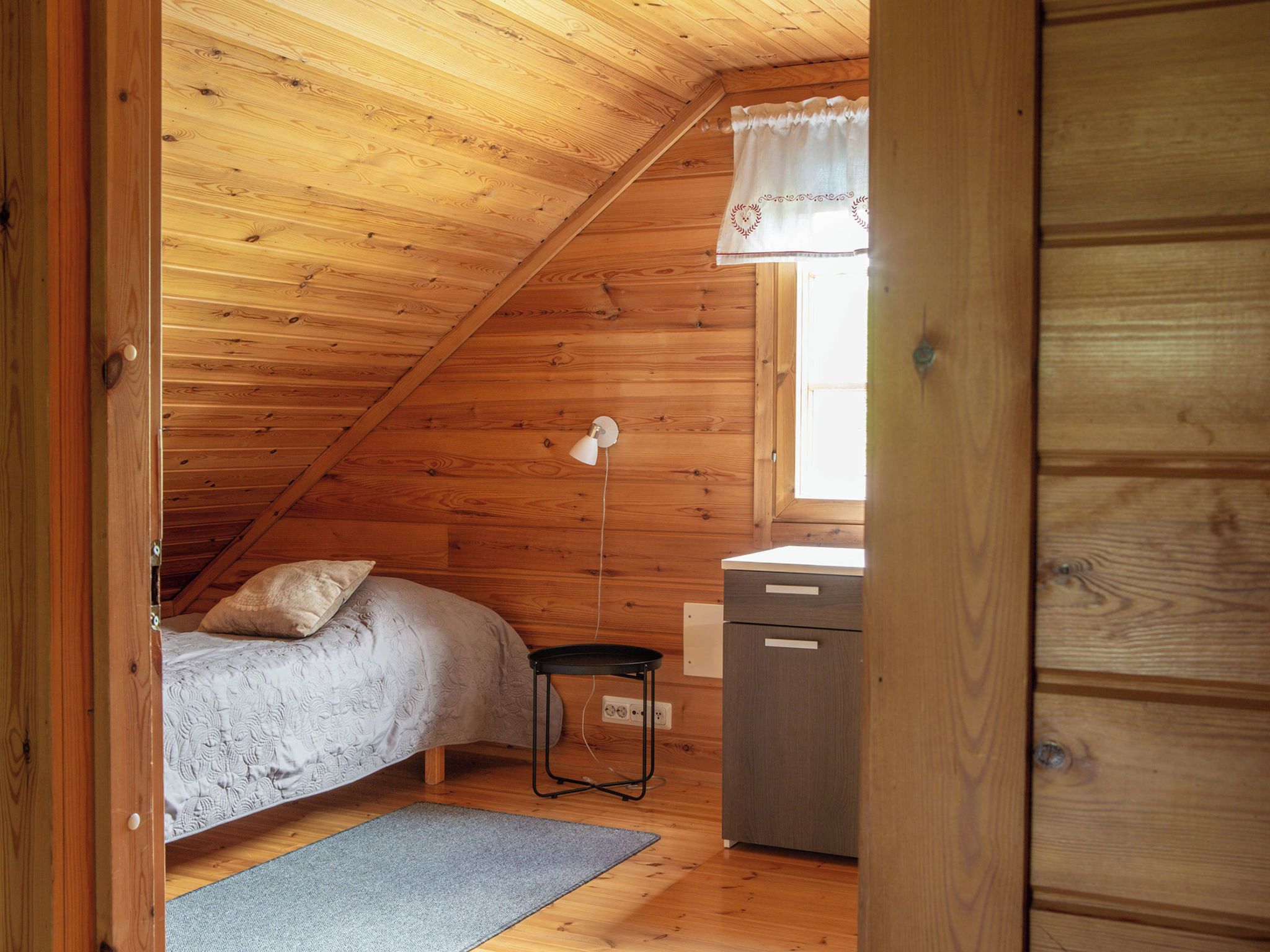 A small, cozy room with wooden walls and a sloped ceiling, featuring a single bed, a small table, and a cabinet.