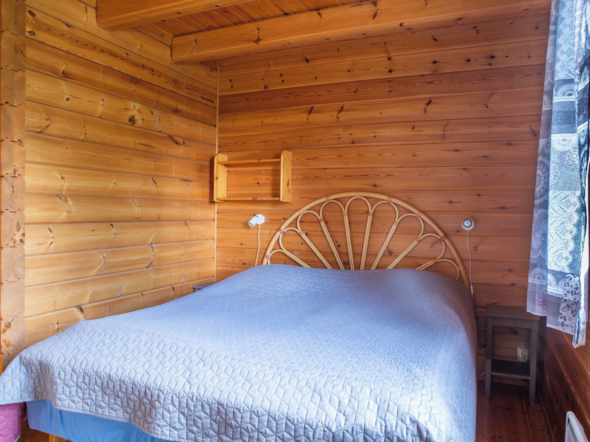 Ein Zimmer in einer Holzhütte mit einem Bett, einem Kopfteil, einem kleinen Tisch und einem Fenster mit Vorhang.