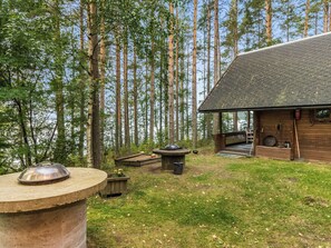 Una cabana rústica amb una zona de barbacoa de pedra, envoltada d'alzines i un cel serè.