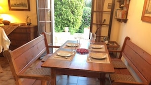 Une table de salle à manger en bois, accompagnée de quatre chaises, mise avec des assiettes et des couverts, dans une pièce dont le sol est carrelé et qui offre une vue sur la verdure à l’extérieur.