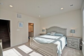 Un dormitorio con una cama, mesitas de noche, un aparador, un tocador con espejo y una puerta que da a otra habitación.