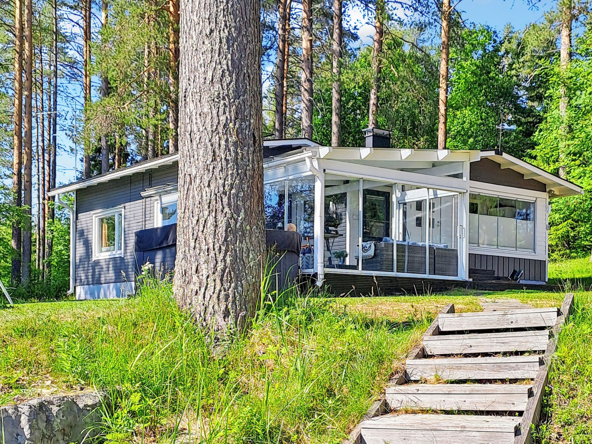 Moderni mökki, jossa on katettu kuisti, ja sen ympärillä on korkeita puita sekä nurmialue.