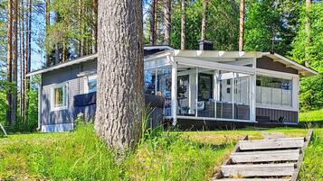 Moderni mökki, jossa on katettu kuisti, ja sen ympärillä on korkeita puita sekä nurmialue.