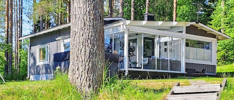 A modern cabin with a covered porch, surrounded by tall trees and a grassy area.