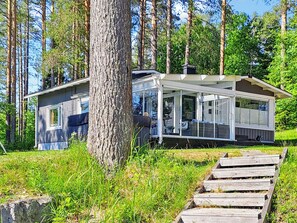 Une cabane moderne dotée d’un porche couvert, entourée de grands arbres et d’une zone herbeuse.