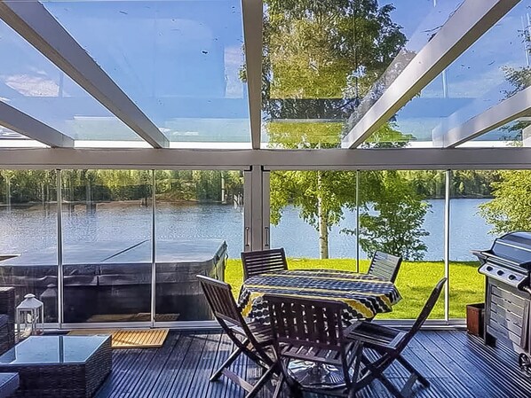 Une terrasse fermée par des baies vitrées, avec une table et des chaises, donnant sur un plan d’eau et une végétation luxuriante.