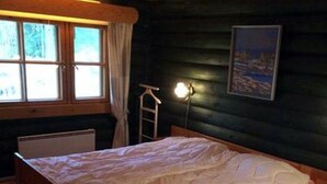 Ett sovrum i en stuga med träpanel, innehållande en säng, ett fönster med gardiner, en stege och en vägglampa.