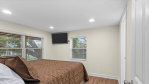 A bedroom with a bed, two windows with blinds, a flat-screen TV mounted on the wall, and a door. - Priya’s Island- sleeps 10 with private dock (Steinhatchee)