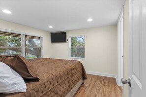 A bedroom with a bed, two windows with blinds, a flat-screen TV mounted on the wall, and a door. - Priya’s Island- sleeps 10 with private dock (Steinhatchee)