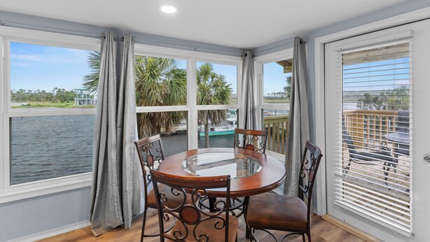 A round glass table with four chairs, large windows with curtains, and a view of a body of water and palm trees. - Priya’s Island- sleeps 10 with private dock (Steinhatchee)