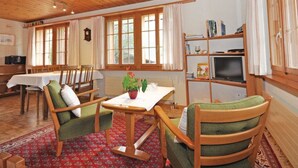 Um quarto aconchegante com assoalho de madeira, uma mesa de jantar com cadeiras, uma estante de livros e uma televisão.