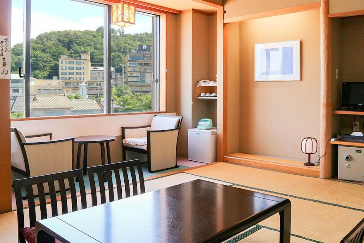 A room with a dining table, chairs, a small refrigerator, a television, and a window with a view of buildings.