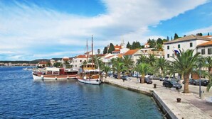 A coastal town with boats docked at a marina, a paved waterfront promenade, and traditional buildings with red-tiled roofs.
