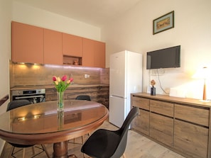 A kitchen with wooden cabinets, a white refrigerator, a glass-top dining table with a chair, and a vase with flowers. - Apartment Rose by Interhome (Brodarica/Žaboric)