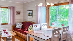 En mysig stuga med en inredning som inkluderar ett matbord i trä, vita stolar, en röd soffa och en utsikt över en skog genom stora fönster.