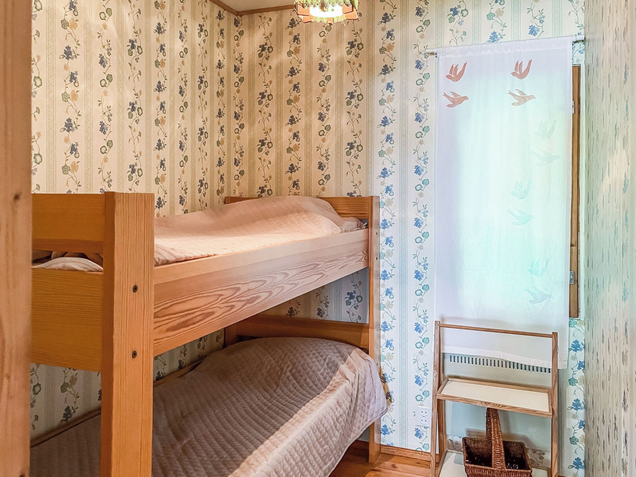 Kerrossängyllä varustettu huone, jossa on kukkatapetti, pieni pöytä ja ikkuna, jossa on verho.