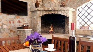 A stone fireplace with a fire, a television mounted above it, and a dining table with chairs and a vase of flowers. - Vila SanAdrija (Trogir)