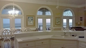 A spacious room with a large white dining table, multiple chairs, and a view of the ocean through arched windows.