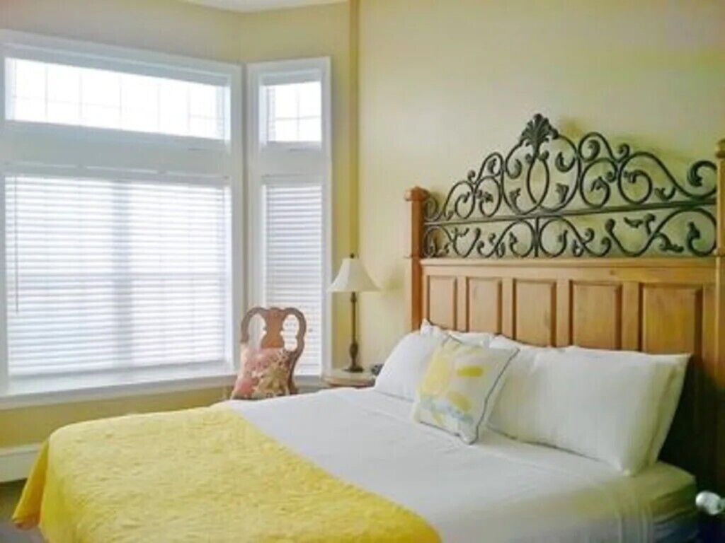 A hotel room with a bed, a window with blinds, a lamp, and a decorative headboard.