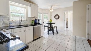 A modern kitchen with white cabinets, a black countertop, and a dishwasher. There is a dining table with chairs and a clock on the wall.