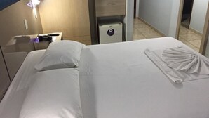 Quarto de hotel com cama bem arrumada, mesa de cabeceira com abajur, gaveteiro e espelho.