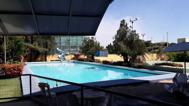 Piscina com área coberta, rodeada por vegetação e um edifício moderno ao fundo.