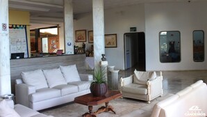 Um hall amplo com um sofá branco, uma mesa de centro de madeira e uma poltrona branca. Há espelhos grandes nas paredes e uma mesa de recepção ao fundo.
