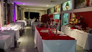 Salão de festas com várias mesas montadas para um evento formal, com toalhas brancas e centros de mesa vermelhos.