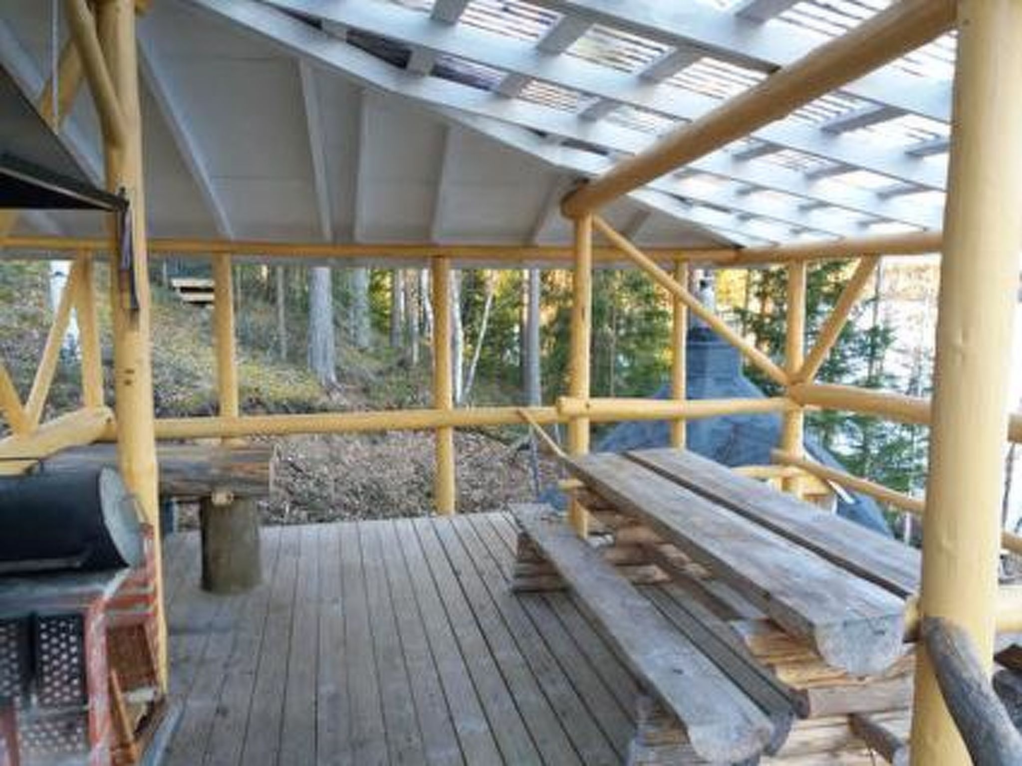 Une terrasse en bois couverte, dotée de bancs et d’une rambarde en métal, entourée d’arbres.