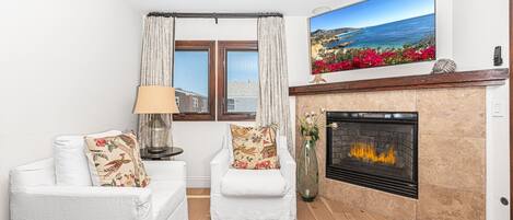 Una sala de estar con chimenea, dos sofás blancos con cojines florales, una mesita de noche con lámpara y un ventanal con cortinas.