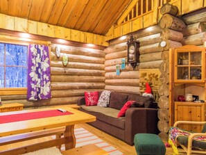 L’intérieur d’un chalet confortable, avec des murs en rondins de bois, une table à manger en bois, un canapé avec des coussins, un fauteuil à bascule et une cheminée en pierre.