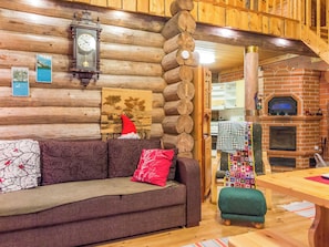 L’intérieur d’une cabane en rondins, avec un mur en bois, une table à manger en bois, un canapé avec des coussins, une cheminée et une horloge.
