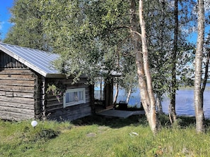 Une cabane rustique avec un toit en métal, entourée d’arbres et bordée par un plan d’eau.