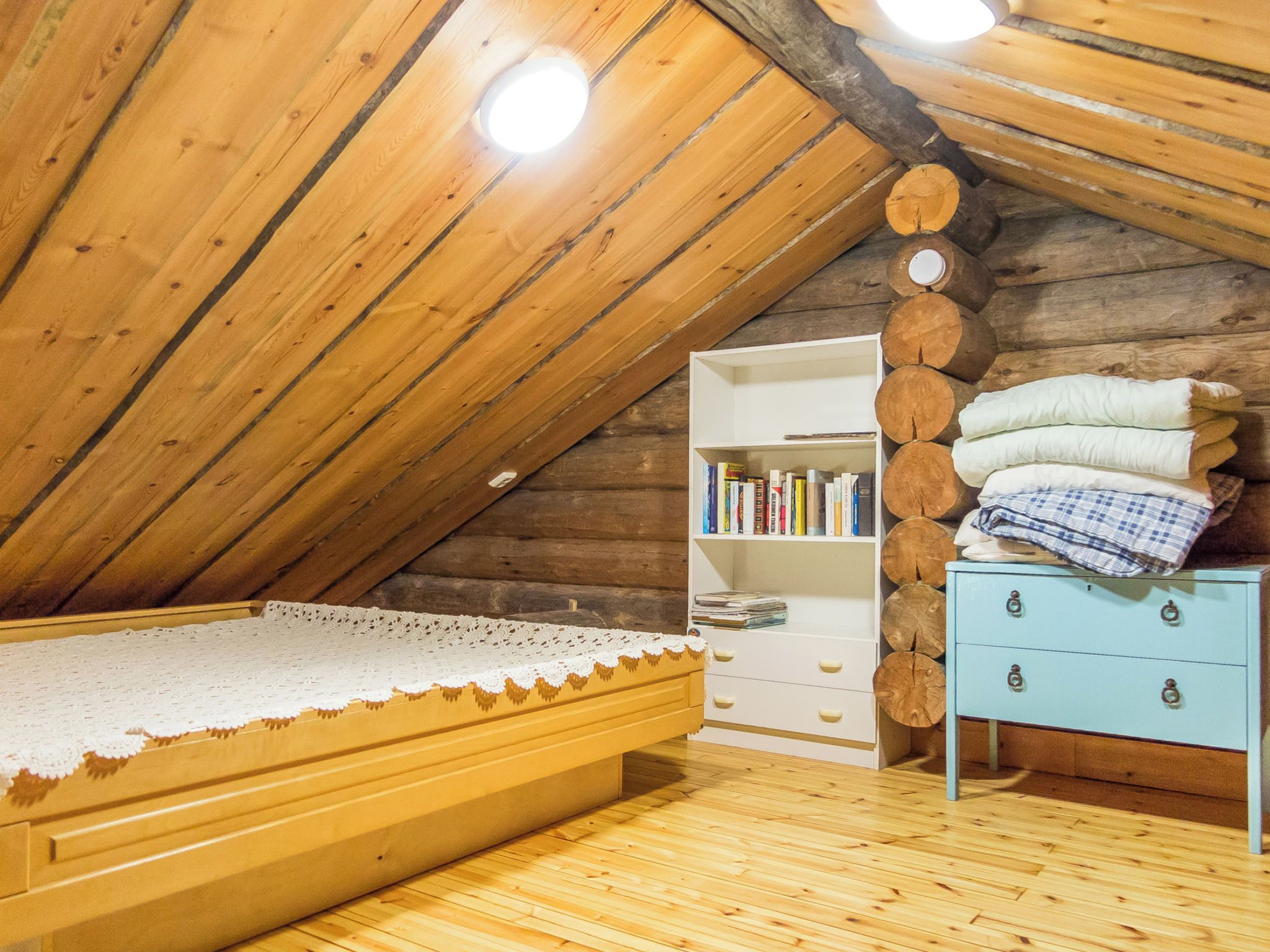 Um quarto no estilo loft com teto inclinado de madeira, uma cama com colcha branca, uma estante branca e um aparador azul.