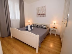 A neatly made bed with pillows and a blanket, two bedside tables with lamps, a painting on the wall, and a door with a radiator. - Apartment Marina by Interhome (Rogoznica/Ražanj)