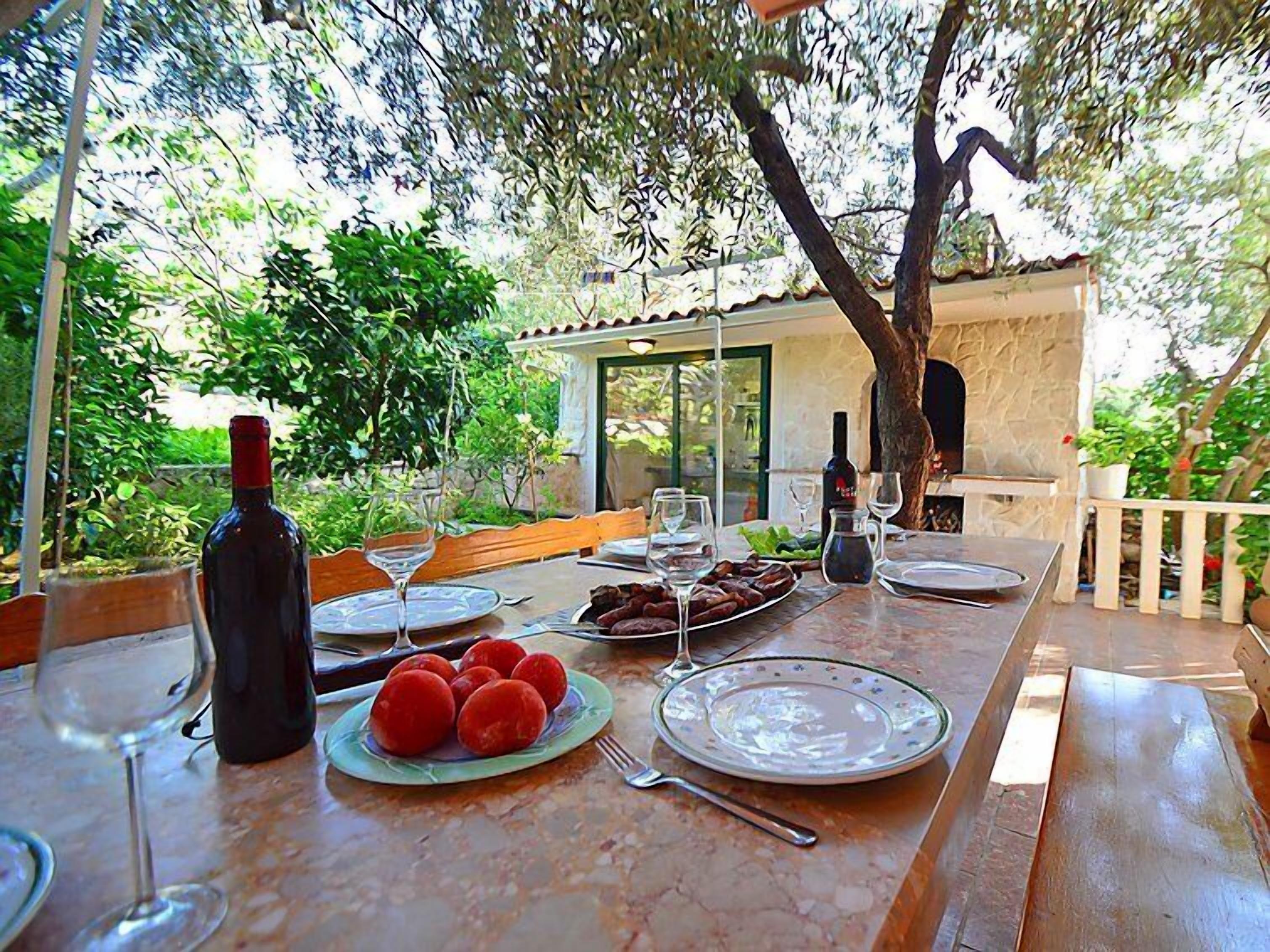An outdoor dining table set with plates, wine glasses, and a bottle, surrounded by greenery and a stone wall.