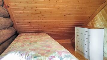 Pieni huone, jossa on yksi sänky, puinen, kalteva katto, valkoinen lipasto laatikoineen ja kukkakuviollinen päiväpeite.